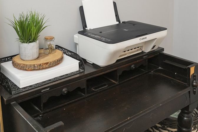 Cozy home office corner: white Canon inkjet printer on a dark vintage wooden desk, decorative tray with stacked paper, wood slice, potted plant and small jar.