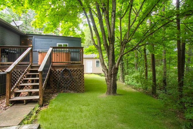Backyard with a raised wooden deck and stairs leading to a hot tub, coiled garden hoses under lattice, a large shade tree on a bright green lawn and dense woods beyond.