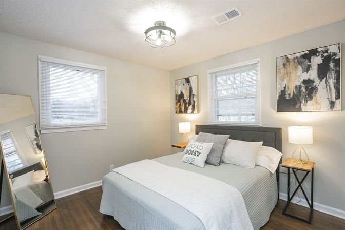 Cozy contemporary bedroom interior with queen bed, gray upholstered headboard and pale blue bedding, two bedside tables with lamps, abstract wall art, leaning full-length mirror, and hardwood floors in soft natural light.