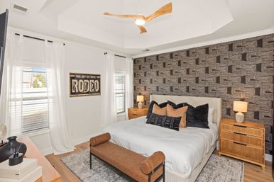 Bright modern master bedroom with geometric textured accent wall, king bed in white bedding with black-and-tan throw pillows, wooden nightstands and lamps, brown upholstered bench at the foot, sheer white curtains and a three-blade wooden ceiling fan.