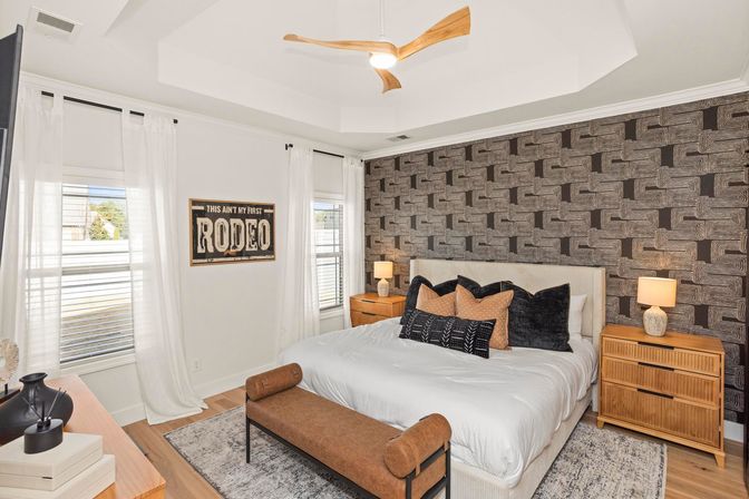 Bright modern master bedroom with geometric textured accent wall, king bed in white bedding with black-and-tan throw pillows, wooden nightstands and lamps, brown upholstered bench at the foot, sheer white curtains and a three-blade wooden ceiling fan.