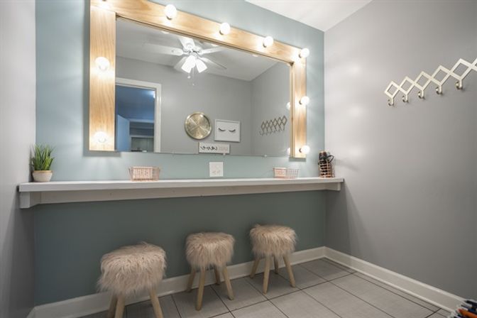 Glam makeup vanity with large wood-framed mirror surrounded by bulbs, long white counter, three fluffy faux-fur stools, gray walls, wall hooks and a small potted plant.