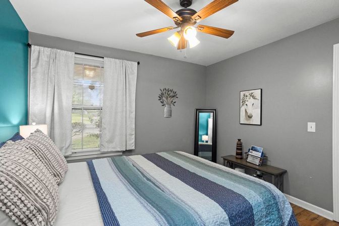 Cozy bedroom with teal accent wall and gray paint, ceiling fan overhead, striped blue quilt and textured pillows on bed, sunlit window with white curtains, full-length mirror and small wooden console table on hardwood floor.