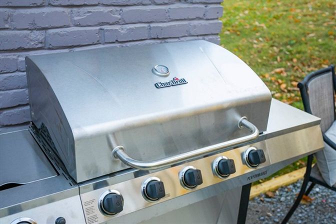 Stainless-steel outdoor gas grill with closed hood, handle and multiple control knobs on a backyard patio beside a painted brick wall and fallen autumn leaves, ready for a cookout.