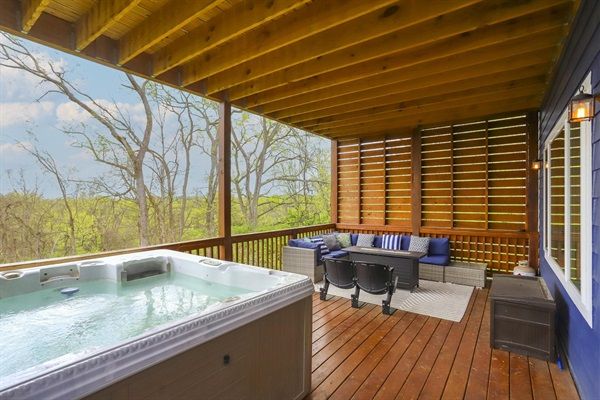 Cozy covered wooden deck patio with bubbling hot tub, cushioned wicker sectional, coffee table and rug, overlooking a green treeline and open sky