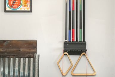 Wall-mounted pool cue rack holding four colorful billiard cues and two wooden triangle racks hanging in a rustic game room corner