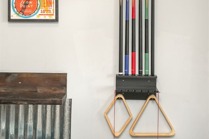 Wall-mounted pool cue rack holding four colorful billiard cues and two wooden triangle racks hanging in a rustic game room corner