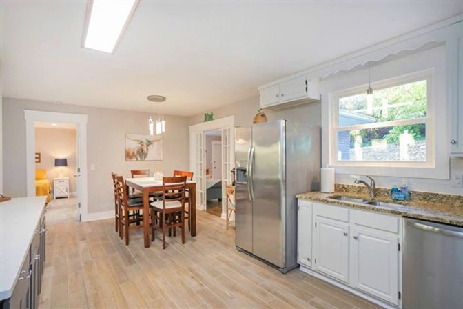 Bright open-plan kitchen and dining room with white cabinets, granite countertops, stainless steel refrigerator, wood-look tile floor and a six-seat wooden table by a garden-facing window