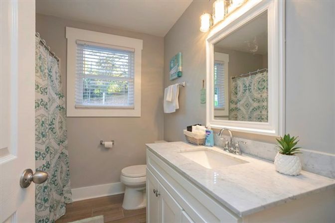 Bright modern small bathroom with marble countertop vanity, white cabinets, undermount sink and framed mirror with vanity lights, toilet, window with blinds, teal patterned shower curtain, wood-look tile floor and small succulent on the counter.