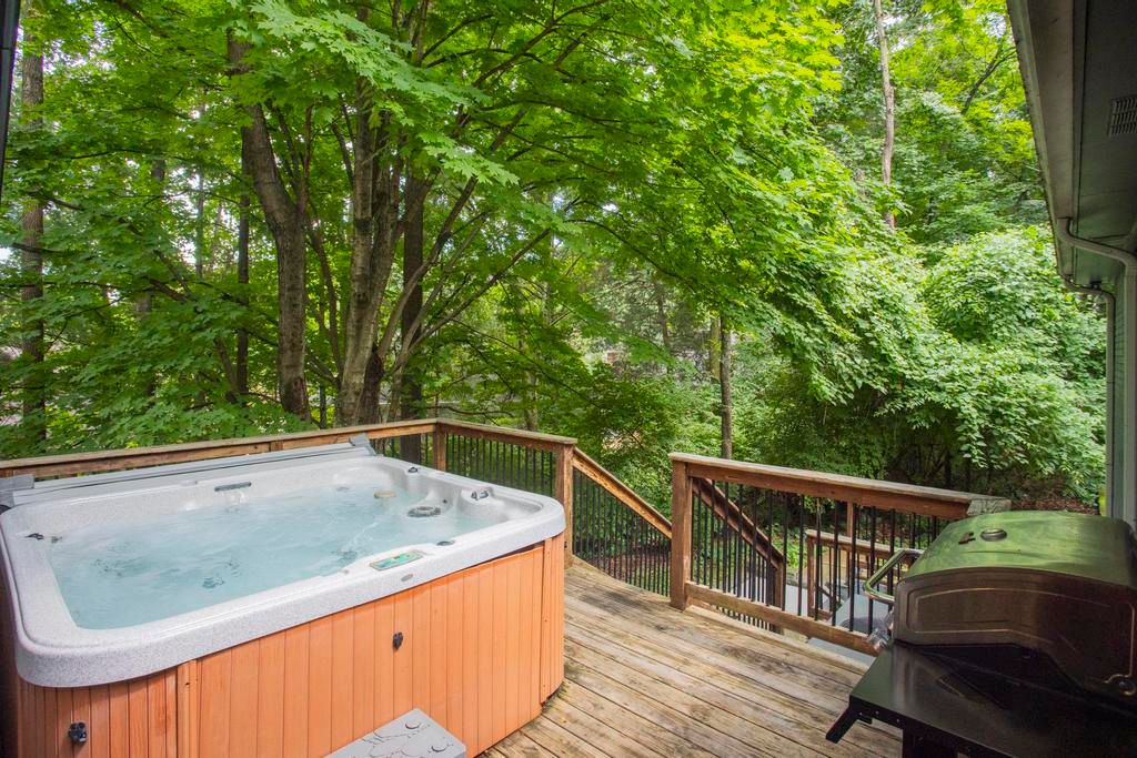 Leafy backyard deck with a bubbling hot tub and outdoor grill overlooking dense green trees and a wooded yard.