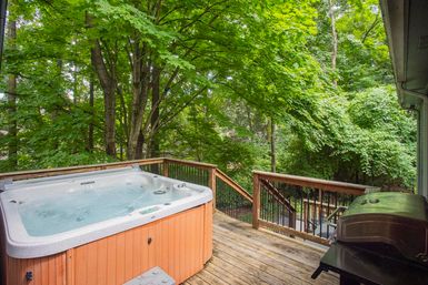 Leafy backyard deck with a bubbling hot tub and outdoor grill overlooking dense green trees and a wooded yard.