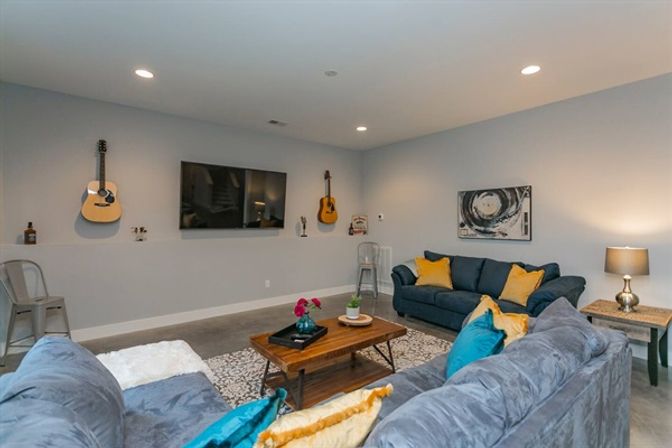 Cozy modern basement family room with gray sectional sofas and blue-and-yellow throw pillows, wall-mounted flat-screen TV flanked by two acoustic guitars, wooden coffee table on a patterned rug, metal stools and a side table lamp.