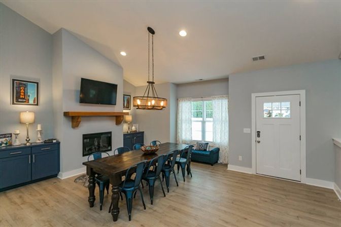 Cozy open-concept dining room in modern farmhouse style with a long dark wood table, eight navy metal chairs, industrial chandelier, navy built-in cabinets and fireplace beneath a wall-mounted TV, window seat and white entry door on light hardwood floors.