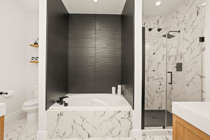 Modern spa-like bathroom featuring a sunken white tub in a black textured-tile alcove, marble-look tile apron and floor, glass-enclosed marble shower with black fixtures, wooden vanity and floating shelves.
