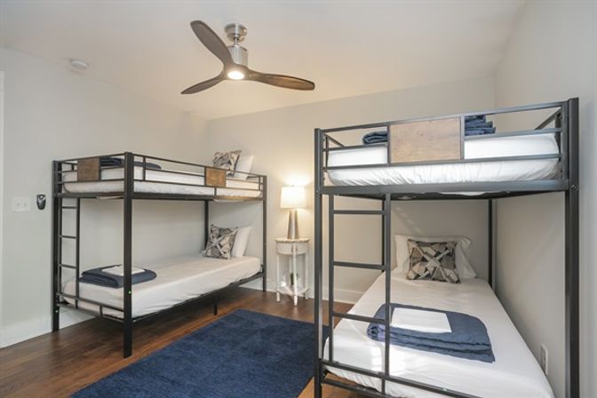 Cozy contemporary guest bedroom with two black metal bunk beds, white linens and patterned pillows, folded navy blankets, hardwood floors, blue area rug, ceiling fan, and a lit bedside lamp against neutral gray walls.