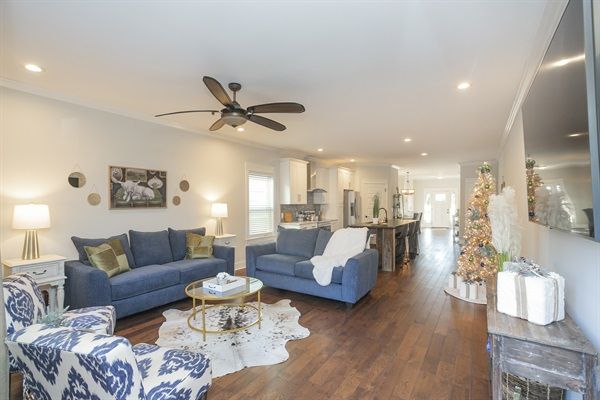 Bright open-concept living room and kitchen with dark hardwood floors, blue sofas, patterned armchair, round gold-accent coffee table, ceiling fan, and decorated Christmas tree by a wall-mounted TV.