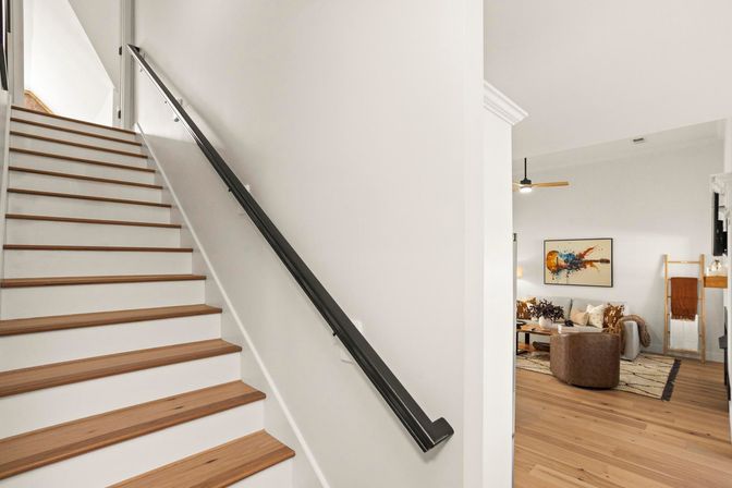 Bright modern home interior: wooden staircase with black handrail leading up, open living room to the right with sofa, colorful wall art and natural wood floors.