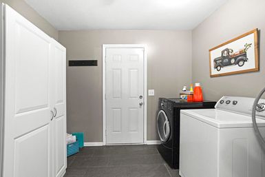 Bright modern laundry room with black front-load washer, white top-load dryer, tall white storage cabinets, gray tile floor, framed vintage pickup art and laundry detergent on the machine.