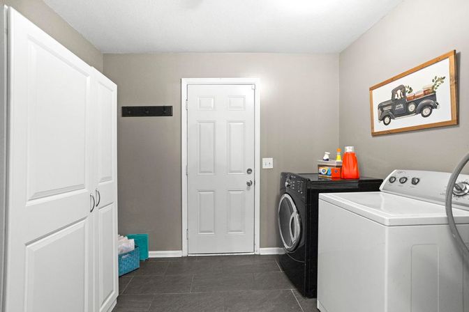 Bright modern laundry room with black front-load washer, white top-load dryer, tall white storage cabinets, gray tile floor, framed vintage pickup art and laundry detergent on the machine.