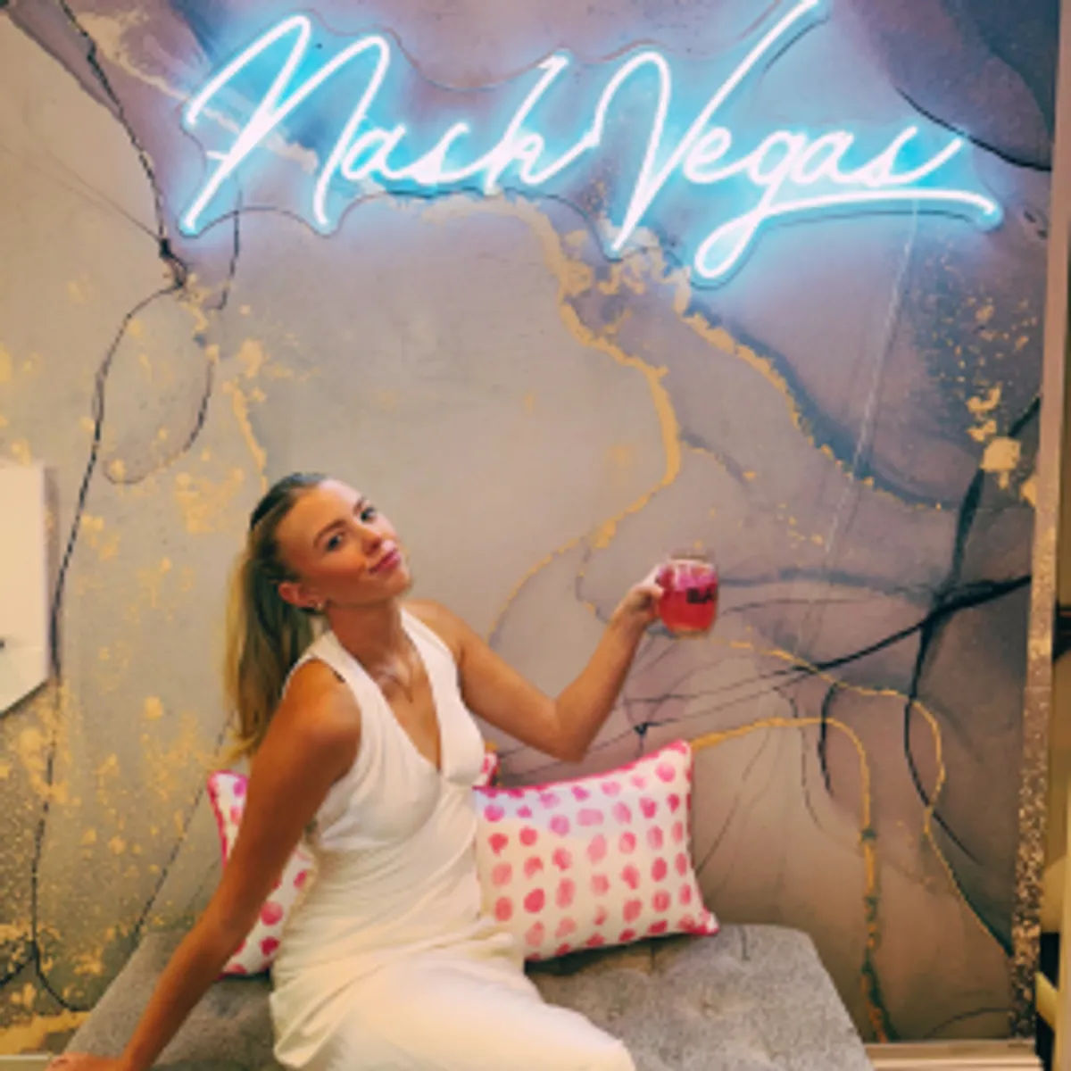 Woman in a white dress lounging on a bench with a pink polka-dot pillow, holding a pink cocktail beneath a blue neon cursive sign on a marble wall with gold accents — trendy indoor lounge backdrop.