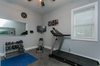 Small residential workout room with treadmill, adjustable bench, dumbbell rack, blue exercise mat, wall mirror, TV, ceiling fan and window blinds