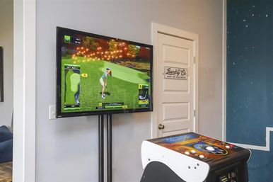 Cozy home game room with wall-mounted TV showing a golf video game and golfer avatar, white arcade cabinet in the foreground, and a nearby door with a laundry sign.