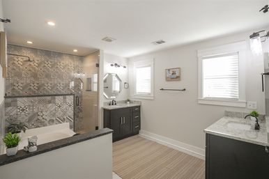 Bright modern residential bathroom with patterned tile walk-in shower, bathtub, dual dark-wood vanities with marble countertops, octagonal mirror and twin windows.