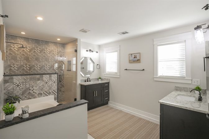 Bright modern residential bathroom with patterned tile walk-in shower, bathtub, dual dark-wood vanities with marble countertops, octagonal mirror and twin windows.