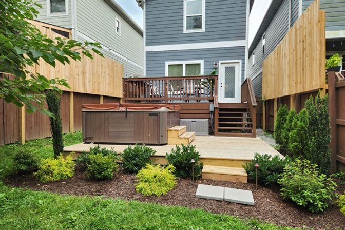 Cozy suburban backyard oasis with a multi-level wooden deck, hot tub on a lower platform, stairs to a raised patio dining area, tall wooden fences and neatly trimmed shrubs.