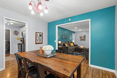 Open-concept dining room with rustic wood table and industrial metal chairs, teal accent wall, and view into living room with gray sectional and yellow pillows.