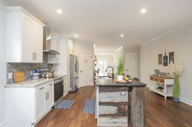Bright open-concept modern farmhouse kitchen with white cabinets, stainless steel appliances, reclaimed-wood island with sink and dark countertop, warm hardwood floors and potted plants.