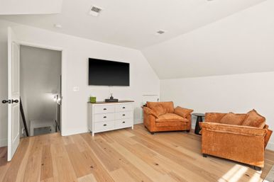 Sunlit upstairs bonus room with light hardwood floors, white sloped ceiling, wall-mounted flat-screen TV above a white six-drawer dresser, and two burnt-orange plush armchairs with a small black side table.
