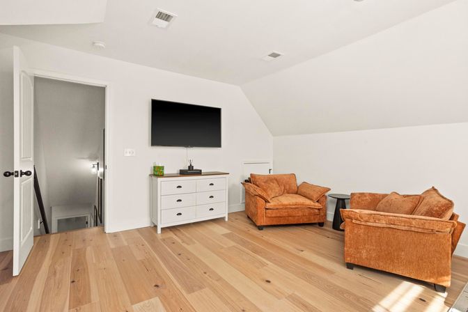 Sunlit upstairs bonus room with light hardwood floors, white sloped ceiling, wall-mounted flat-screen TV above a white six-drawer dresser, and two burnt-orange plush armchairs with a small black side table.