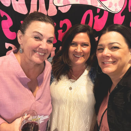 Three smiling women friends taking a close-up group selfie at a casual gathering, one holding a glass of red wine, in front of a bold pink and black patterned backdrop.