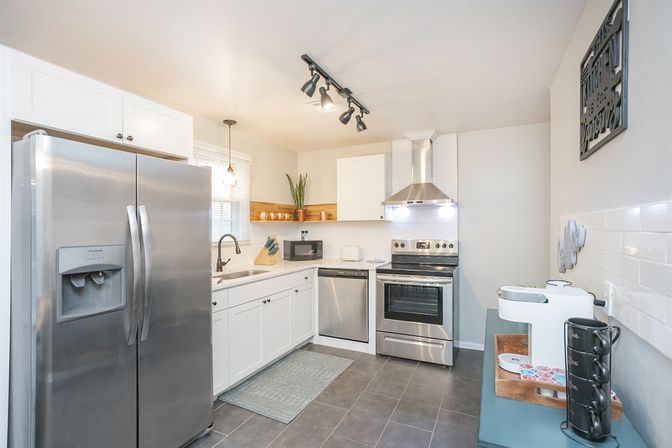Bright modern kitchen with stainless steel fridge, oven and dishwasher, white shaker cabinets, subway tile backsplash, farmhouse sink, pendant and track lighting, gray tile floor and a coffee station.