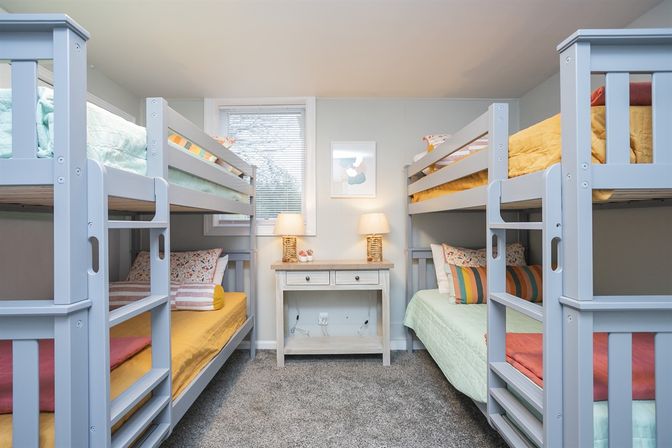 Cozy family bunk bedroom with two light-gray twin bunk beds, colorful yellow and mint bedding, central window, wooden nightstand with two lamps, and plush carpet.