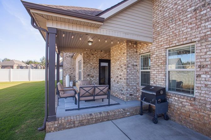Cozy covered brick patio with outdoor seating, string lights and a charcoal grill overlooking a fenced green lawn in a suburban backyard.