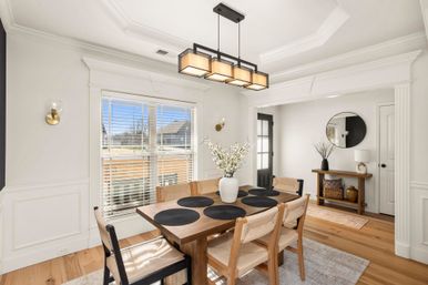 Sunlit modern dining room with a wooden table and six woven chairs, black rectangular pendant light, white vase of branches atop black round placemats, large window with blinds, white wainscoting, hardwood floors and an entryway console with round mirror.