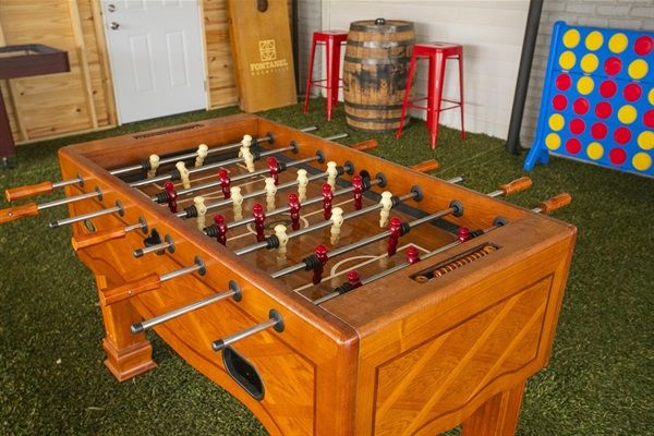 Wooden foosball table on green artificial-turf floor in a casual indoor game room with a giant Connect Four, cornhole board, wooden barrel and red metal stools