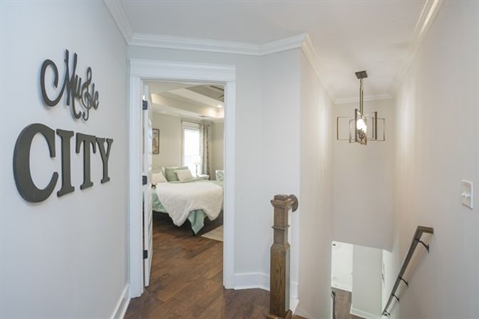 Upstairs hallway with dark hardwood floors, decorative wall letters reading 'CITY', white-trimmed doorway into a bright bedroom with mint-green bedding, wooden banister and modern pendant light over the stairwell.