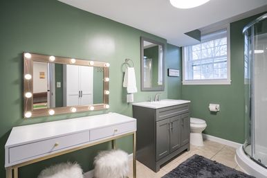 Cozy modern bathroom with green walls, gray sink vanity, white makeup table with Hollywood-style lighted mirror, corner glass shower, toilet, window, and fluffy stools.