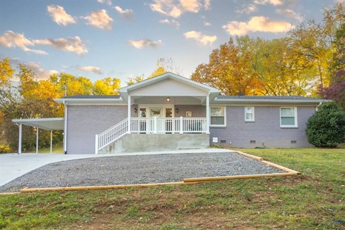 Suburban single-story brick ranch house with covered front porch and white railings, adjacent carport, gravel parking pad and autumn-colored trees under a golden sky.