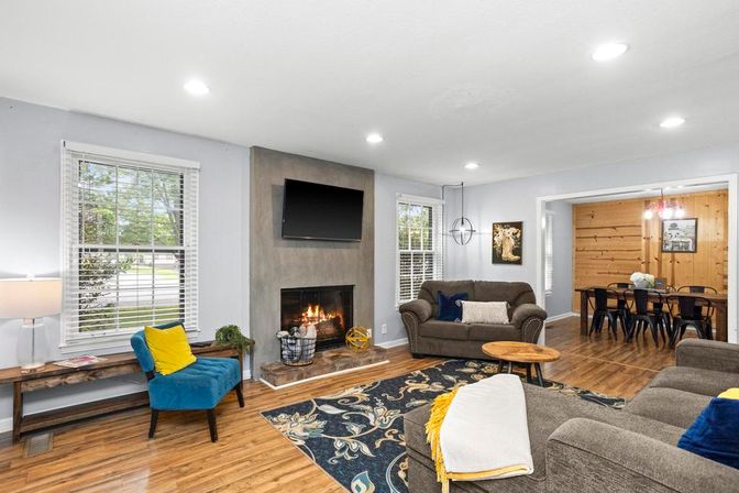 Cozy open-concept living room with hardwood floors, modern fireplace and wall-mounted TV, gray sofas, blue accent chair with yellow pillow, patterned area rug, and adjoining wood-paneled dining area.