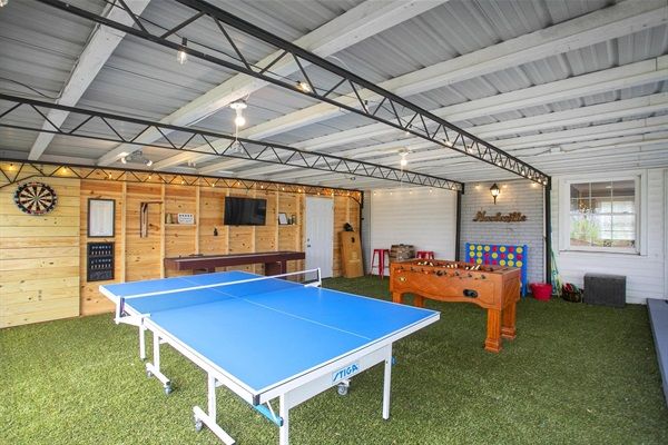 Bright covered backyard game room with blue ping pong table, wooden foosball table, dartboard, wall-mounted TV, and artificial turf flooring under exposed metal beams.