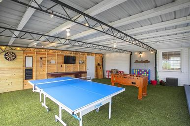 Bright covered backyard game room with blue ping pong table, wooden foosball table, dartboard, wall-mounted TV, and artificial turf flooring under exposed metal beams.