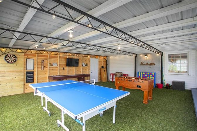 Bright covered backyard game room with blue ping pong table, wooden foosball table, dartboard, wall-mounted TV, and artificial turf flooring under exposed metal beams.
