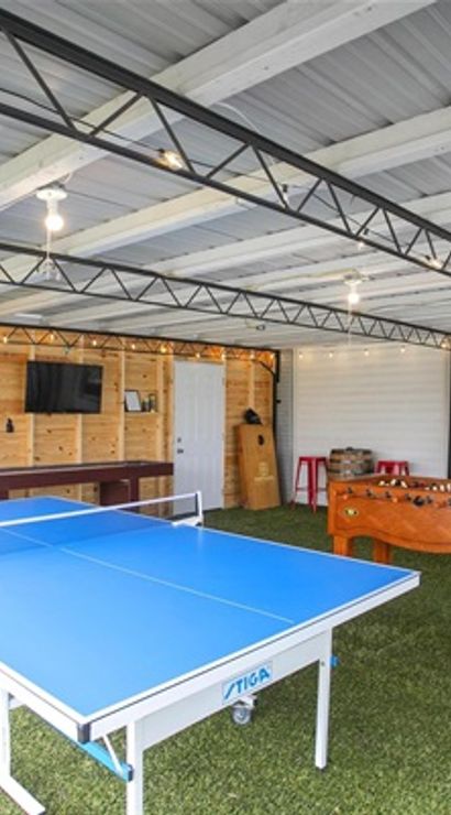Bright covered backyard game room with blue ping pong table, wooden foosball table, dartboard, wall-mounted TV, and artificial turf flooring under exposed metal beams.