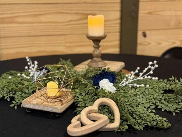 Rustic wedding table centerpiece on black tablecloth with interlocking wooden hearts, faux greenery and blue-white flowers, geometric gold tealight holder with LED candle on a wooden block, and a taller wooden candlestick with glowing candle.