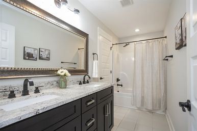 Bright modern master bathroom with double-sink granite countertop, dark wood vanity, large framed mirror, matte black faucets, tiled floor and bathtub with neutral shower curtain