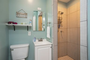 Compact teal-painted bathroom interior with a white toilet and vanity, wall shelf with toiletries, mirror and towel ring, and a beige-tiled walk-in shower with chrome fixtures.